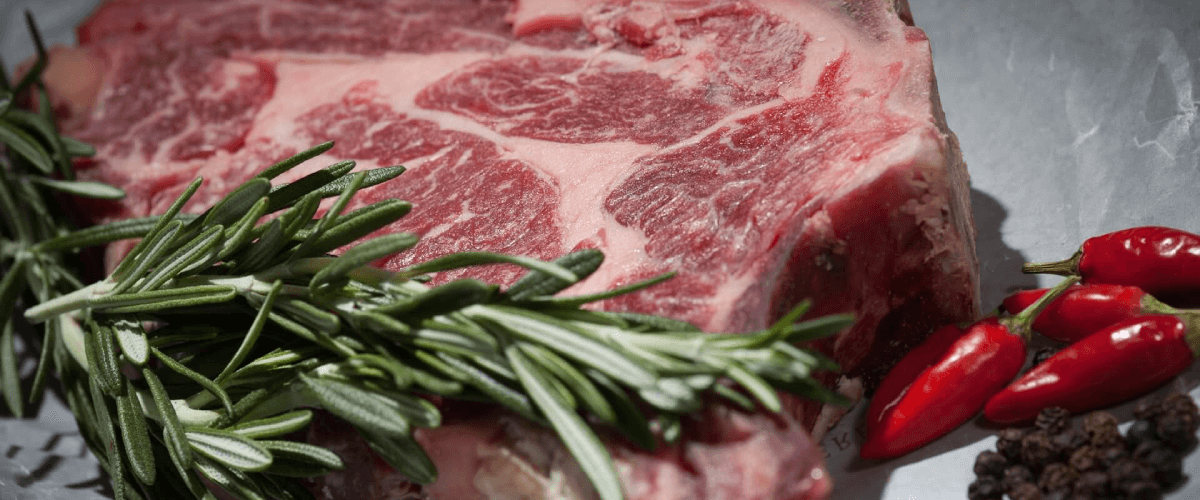 Una bistecca alla fiorentina cruda, pronta per essere cucinata. La bistecca è un taglio spesso di carne di manzo con un osso a T al centro, caratteristica della cucina toscana. La carne è di colore rosso intenso e presenta venature bianche di grasso. La bistecca viene tradizionalmente cotta alla griglia, lasciando l'interno rosato e succoso. Questa immagine mostra la bistecca cruda, prima di essere cotta, evidenziando la qualità e la freschezza del taglio di carne.